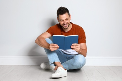 Handsome man reading book on wooden floor near light wall