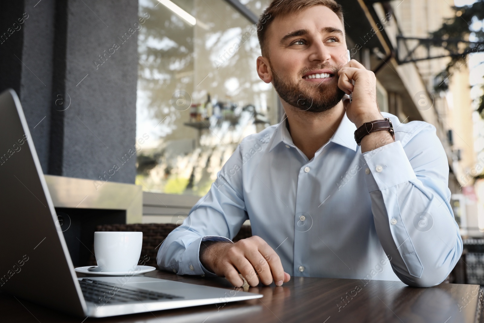 Image of Happy young man with laptop talking on phone at outdoor cafe