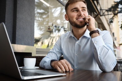 Image of Happy young man with laptop talking on phone at outdoor cafe