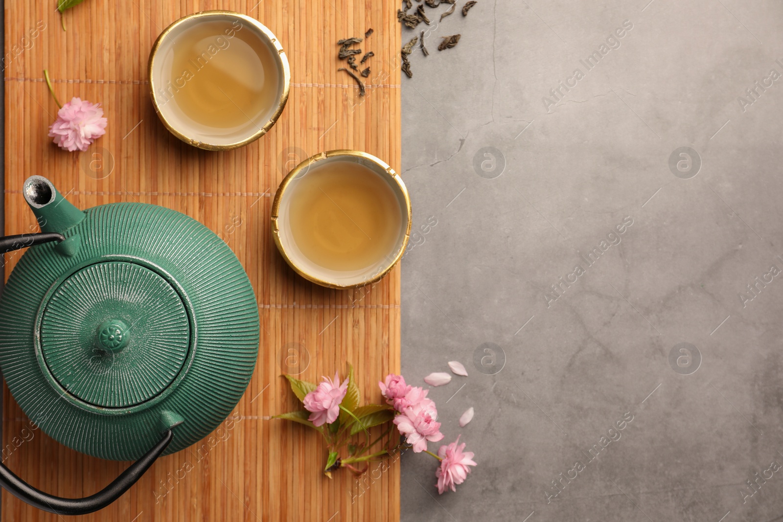 Photo of Traditional ceremony. Cups of brewed tea, teapot and sakura flowers on grey table, flat lay with space for text