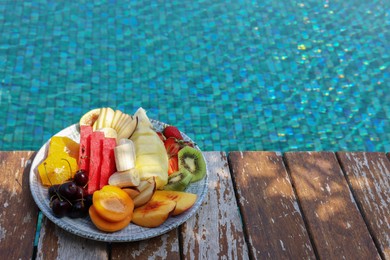 Photo of Plate with fresh fruits on wooden deck near outdoor swimming pool, space for text. Luxury resort