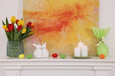 Photo of Easter decorations. Bouquet of tulips in vase, burning candles and bunny figures on fireplace at home