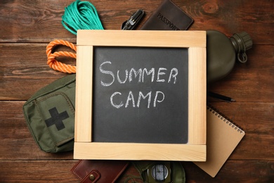 Chalkboard with text SUMMER CAMP and camping equipment on wooden background, flat lay