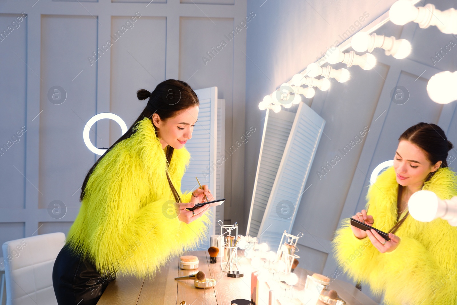 Photo of Young woman applying make up near illuminated mirror indoors