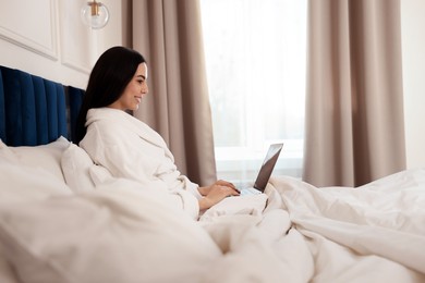 Photo of Happy young woman working with laptop on bed in hotel room