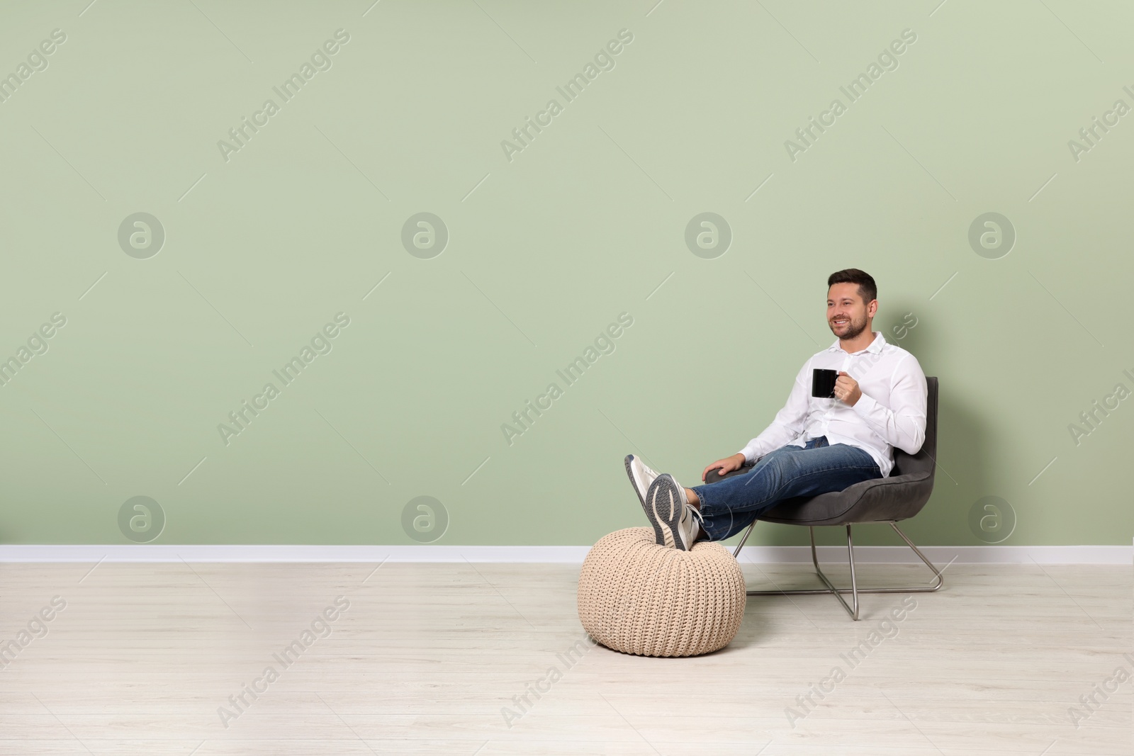 Photo of Happy man with cup of drink sitting in armchair near light green wall indoors, space for text