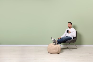 Photo of Happy man with cup of drink sitting in armchair near light green wall indoors, space for text