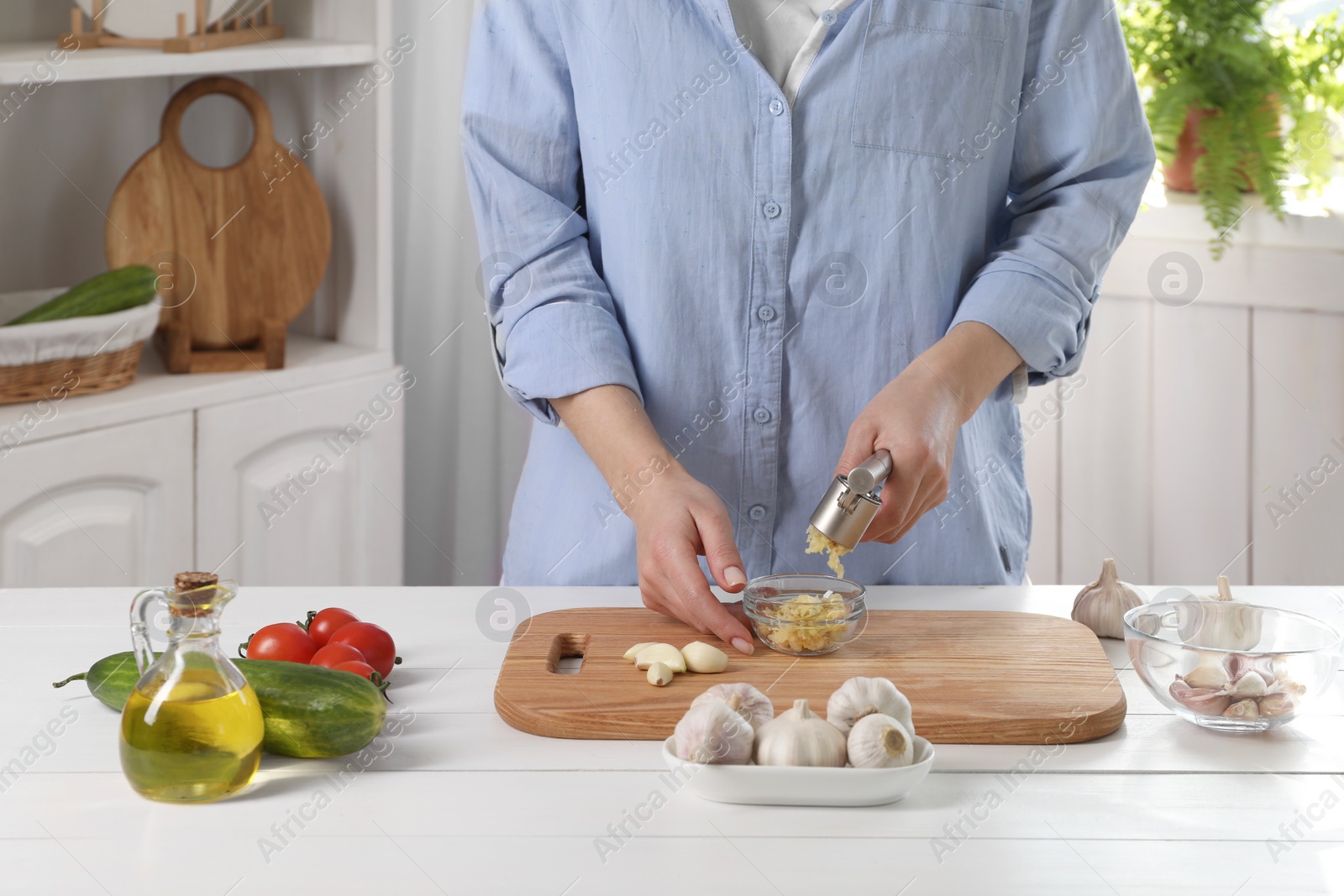 Photo of Woman squeezing garlic with press at white wooden table in kitchen, closeup