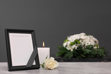 Photo frame with black ribbon, burning candle, rose on light table and wreath of flowers near grey wall indoors, space for text. Funeral attributes