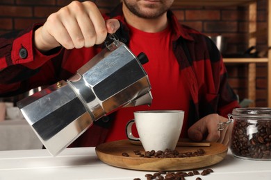 Man pouring aromatic coffee from moka pot into cup at white wooden table indoors, closeup