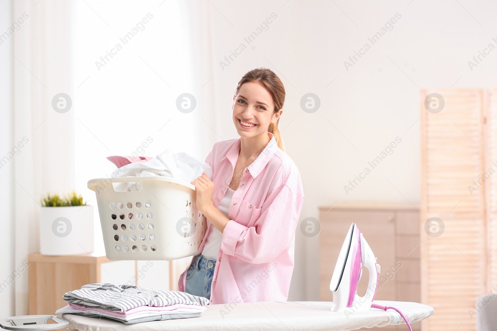 Photo of Young pretty woman holding basket of clean laundry at ironing board indoors