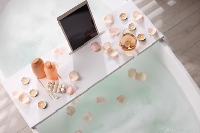 Photo of Wooden tray with tablet, wine, toiletries and flower petals on bathtub in bathroom, above view