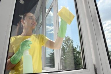 Happy young woman cleaning window glass with sponge cloth and spray indoors