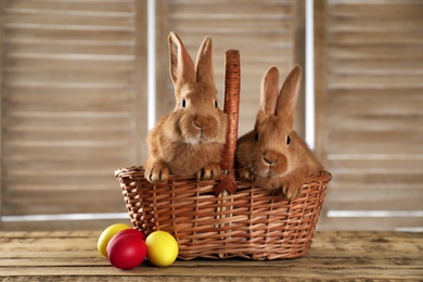 Photo of Cute bunnies, basket and Easter eggs on wooden table against blurred background