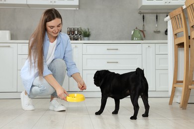 Beautiful young woman feeding her adorable Pug dog in kitchen