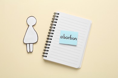 Photo of Notebook with word Abortion and pregnant woman paper cutout on beige background, flat lay