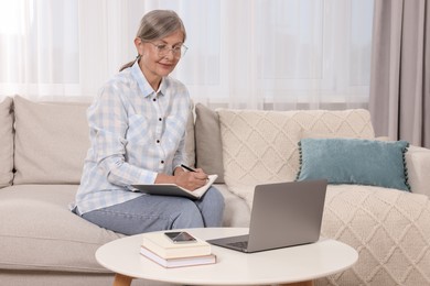 Photo of Beautiful senior woman writing something in notebook while working with laptop at home