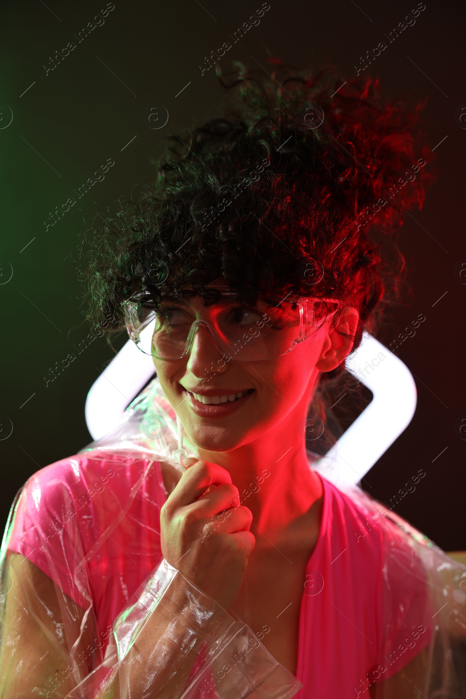 Photo of Beautiful young woman in transparent coat and sunglasses with square lamp on color background in neon lights