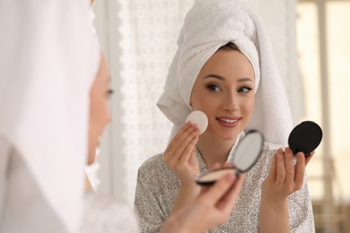 Beautiful young woman applying face powder with puff applicator in front of mirror at home