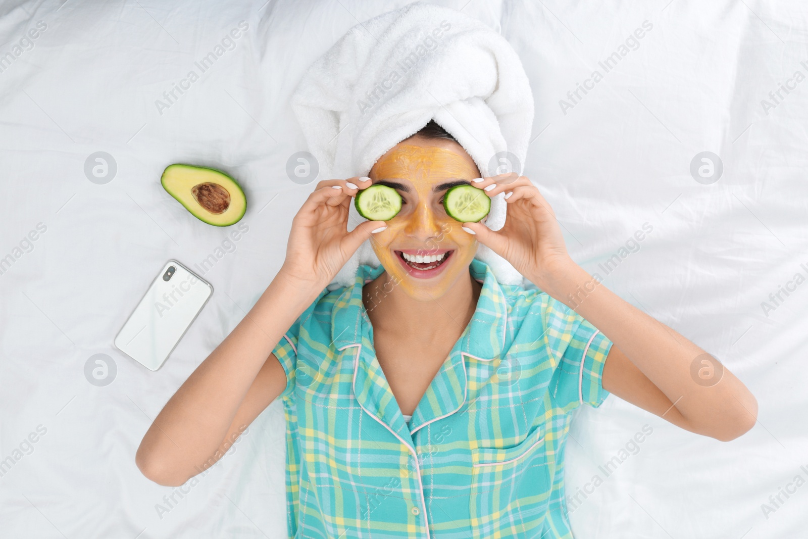 Photo of Young woman with facial mask and cucumber slices lying on bed, top view