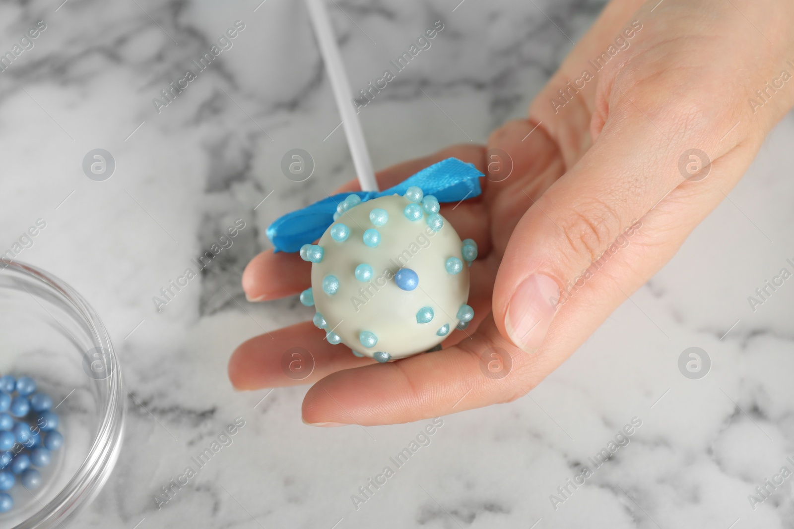 Photo of Young woman holding tasty cake pop with blue sprinkles above white marble table, closeup