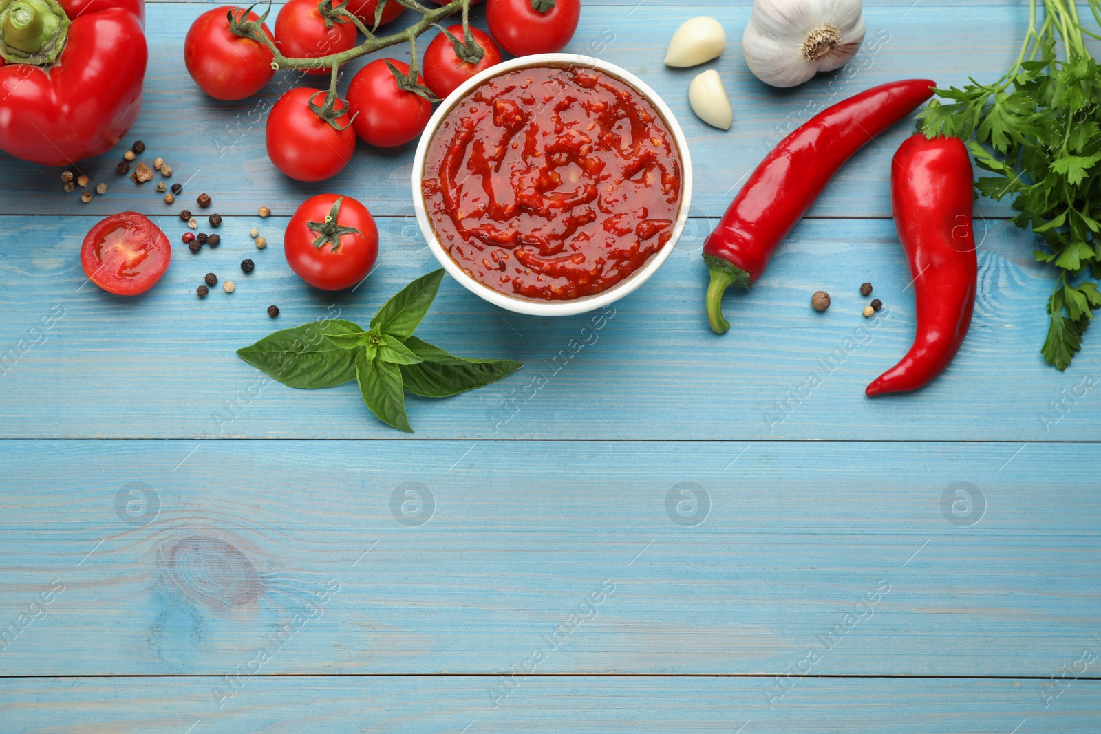 Photo of Delicious adjika sauce in bowl and ingredients on light blue wooden table, flat lay. Space for text