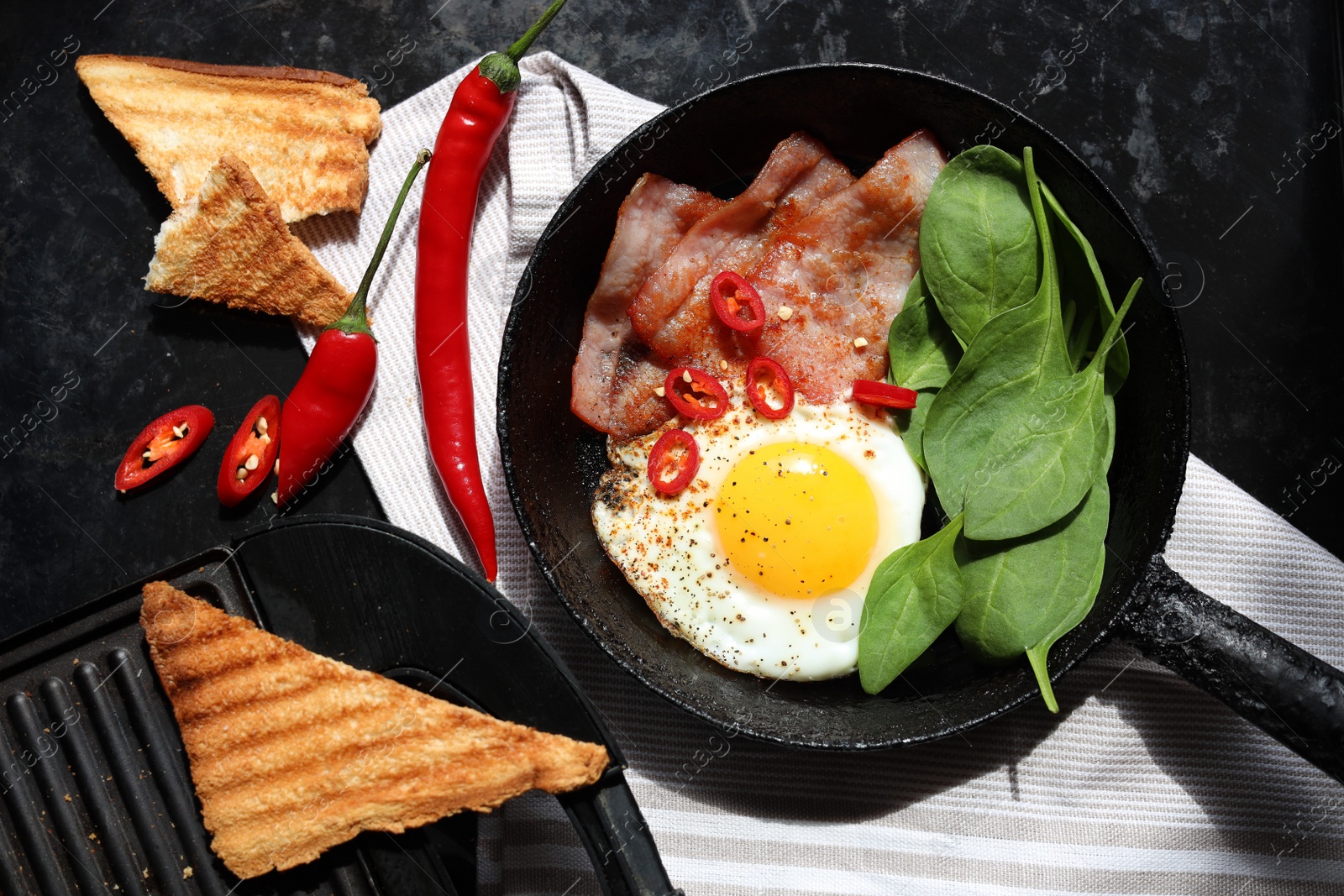 Photo of Tasty fried egg with bacon, chili pepper and spinach on black table, flat lay