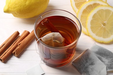 Photo of Tea bags, glass of hot drink, cinnamon sticks and lemons on white wooden table, closeup