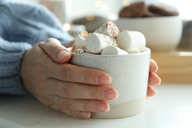 Woman holding cup of delicious hot cocoa with marshmallows at white table indoors, closeup. Winter drink
