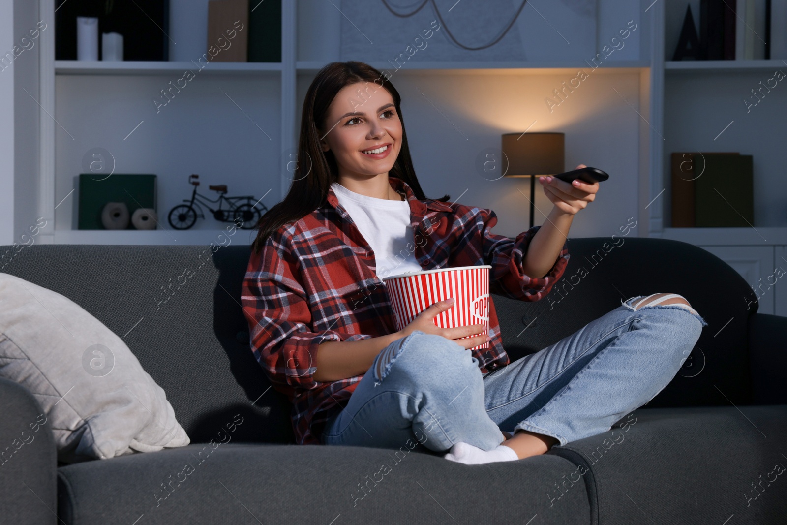 Photo of Happy woman holding popcorn bucket and changing TV channels with remote control at home in evening