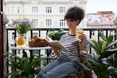 Young woman with refreshing drink reading book near beautiful houseplants on balcony