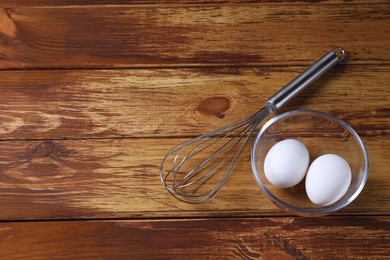 Metal whisk and eggs in bowl on wooden table, flat lay. Space for text