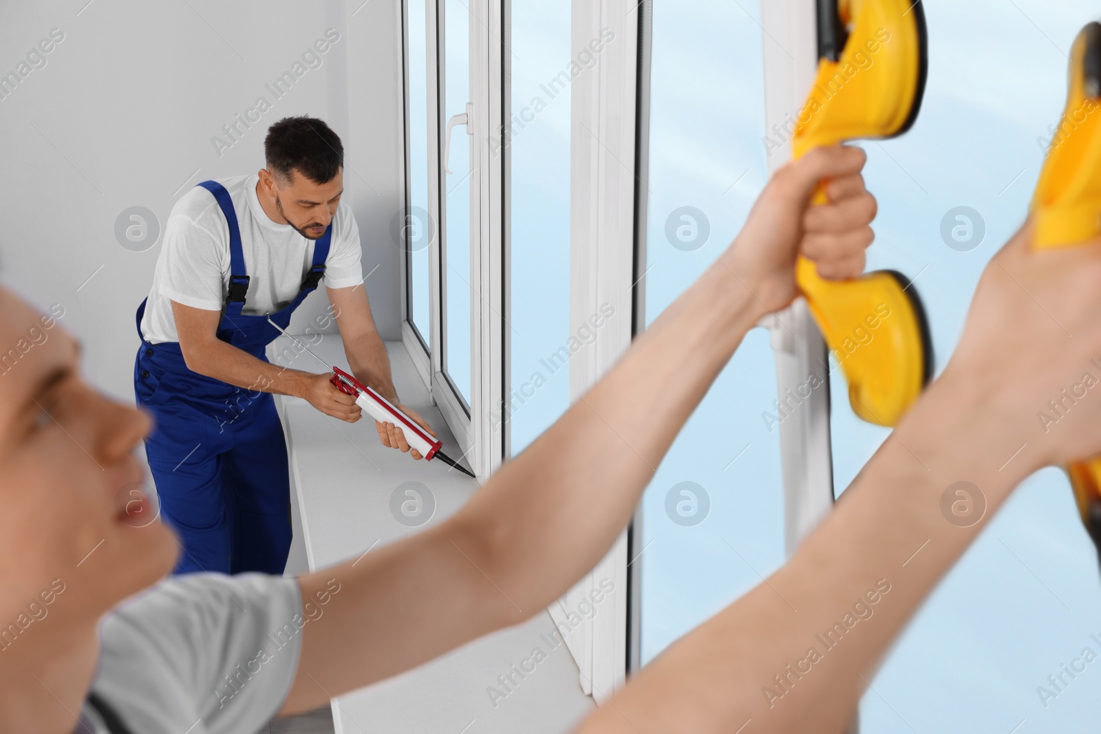 Photo of Workers with tube of sealant and suction lifters installing plastic windows indoors