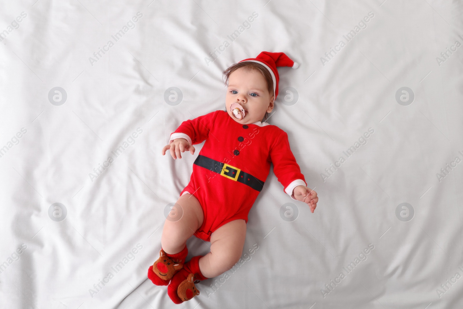 Photo of Cute baby wearing festive Christmas costume with pacifier on white bedsheet, top view