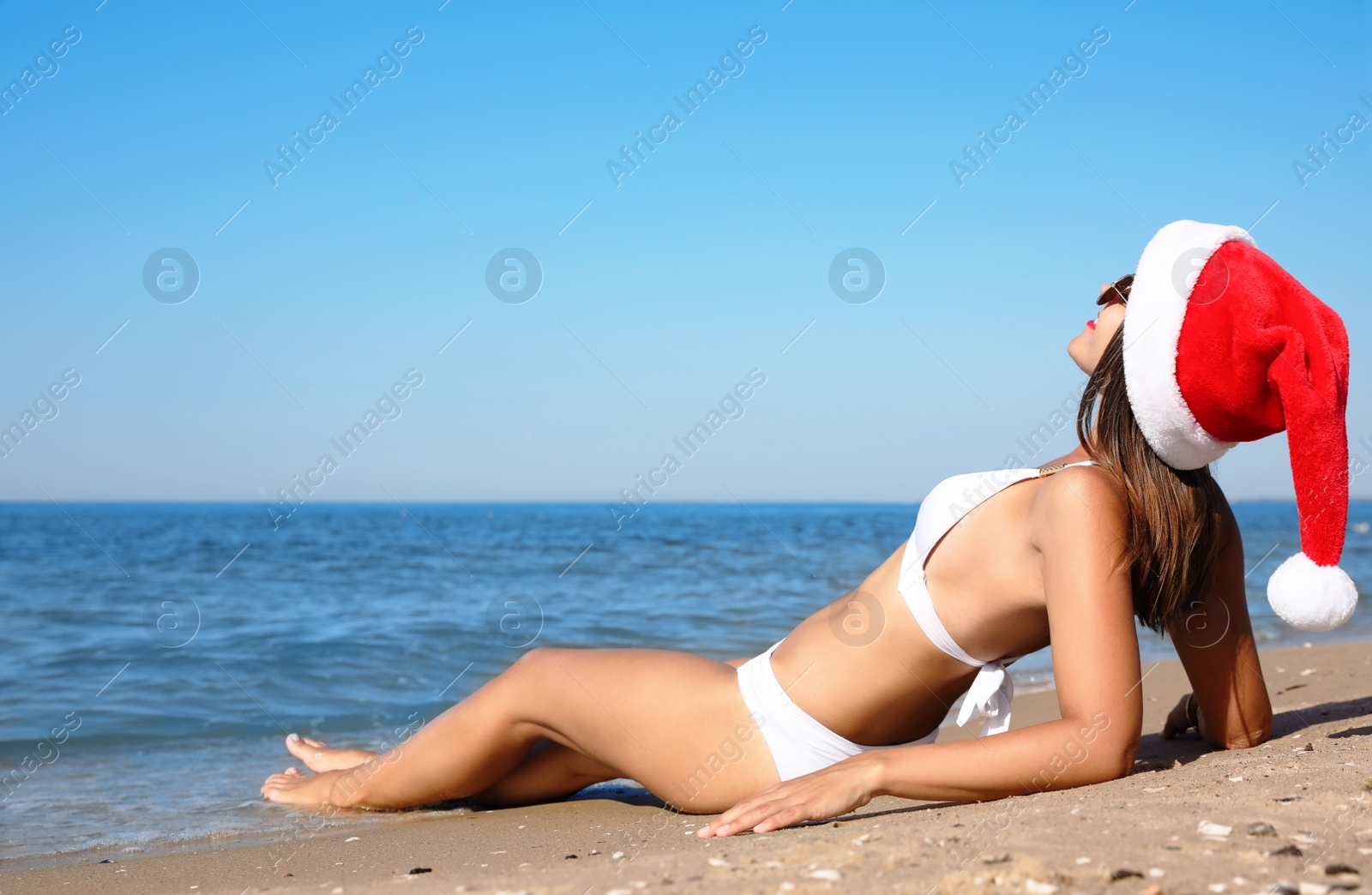 Photo of Young woman wearing Santa hat and bikini on beach, space for text. Christmas vacation