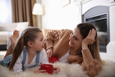 Photo of Happy woman and her daughter with cups of hot drink resting near fireplace at home