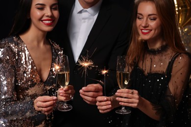 Photo of Happy friends celebrating New Year against black background, focus on hands with glasses of sparkling wine and sparklers