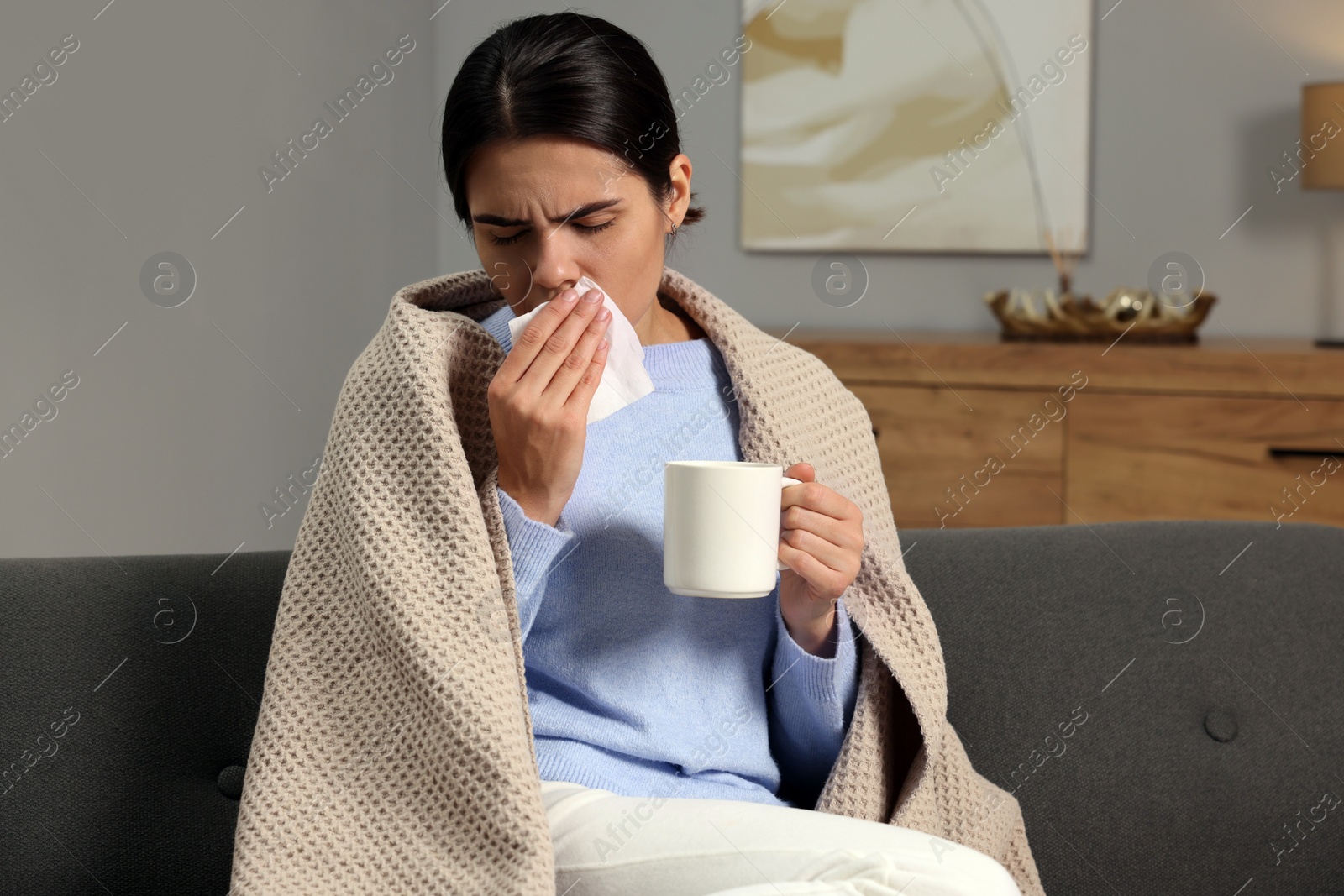 Photo of Woman with tissue and cup of drink coughing on sofa at home. Cold symptoms