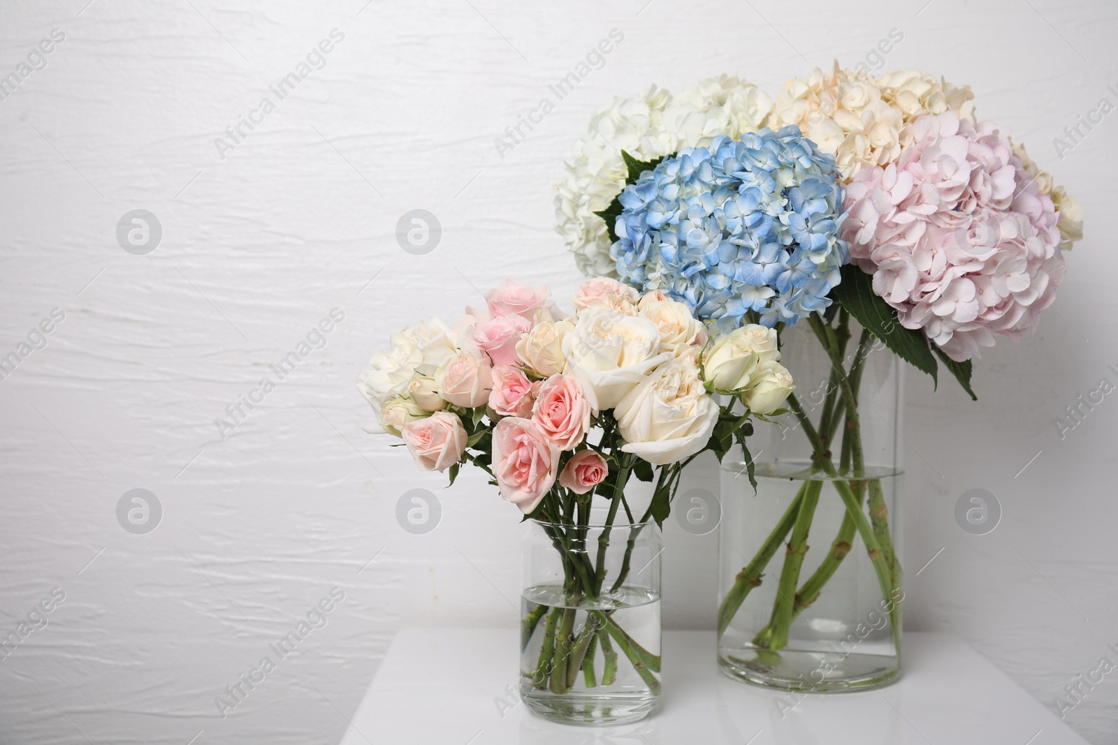 Photo of Beautiful hydrangea and rose flowers in vases on white bedside table indoors, space for text