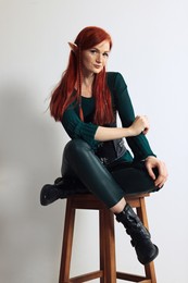 Beautiful redhead elf girl sitting on stool against light background