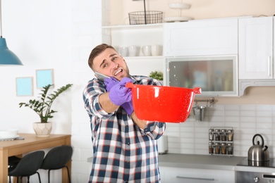 Photo of Emotional young man calling plumber while collecting water leakage from ceiling in kitchen