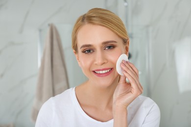 Happy young woman cleaning face with cotton pad in bathroom