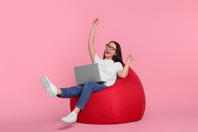 Photo of Happy young woman with laptop sitting on beanbag chair against pink background