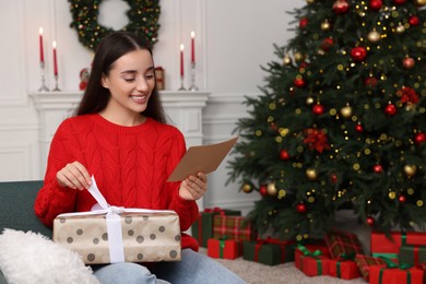 Happy young woman with Christmas gift reading greeting card at home