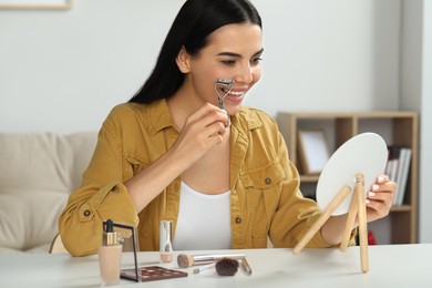 Photo of Beautiful young woman using eyelash curler while doing makeup at table indoors