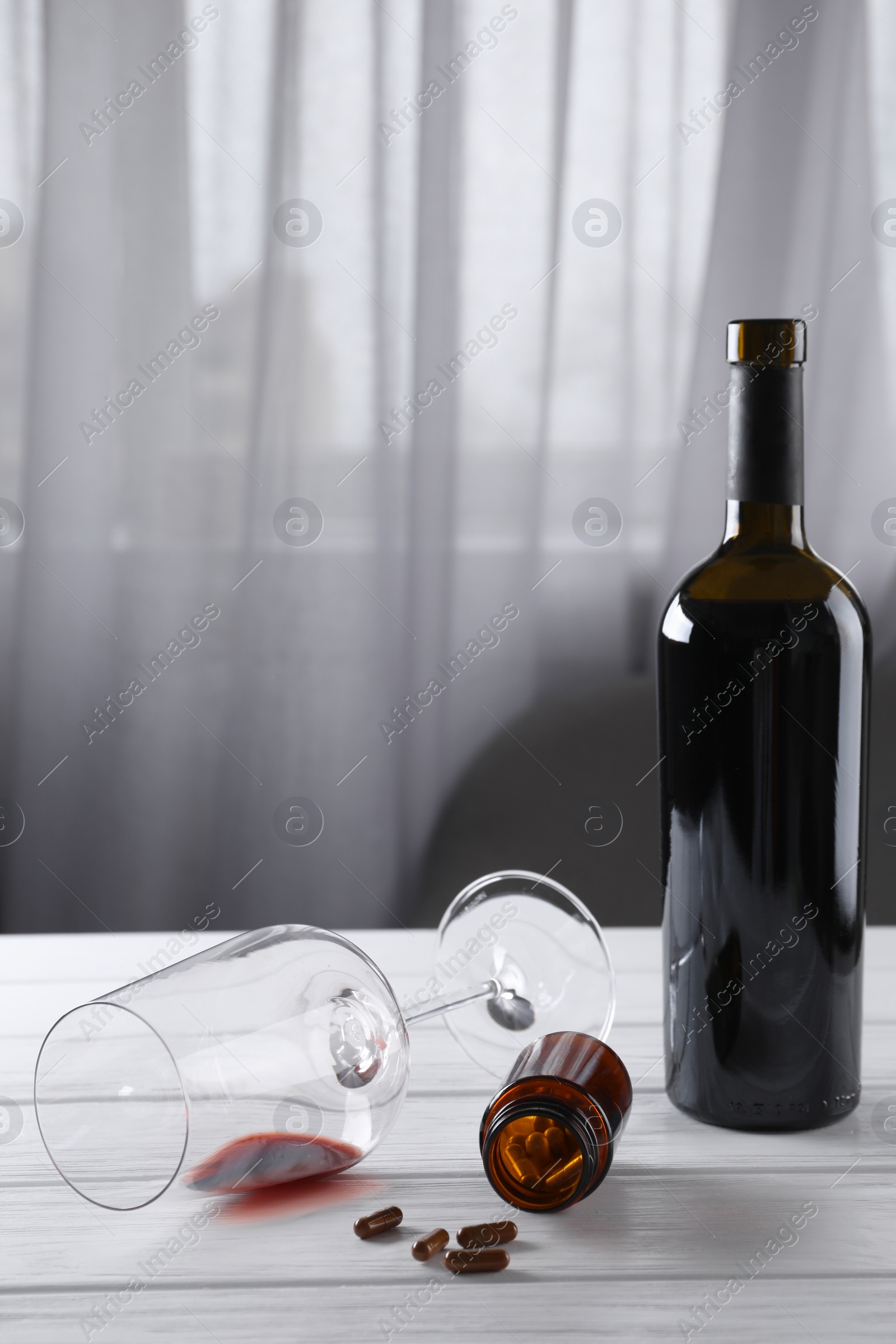 Photo of Alcohol and drug addiction. Overturned glass with red wine, bottle and pills on white wooden table