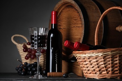 Photo of Glass of red wine, bottles, wicker basket, grapes and wooden barrel on black table