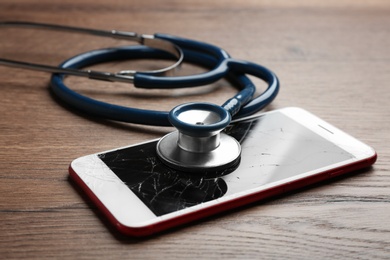 Modern smartphone with broken display and stethoscope on wooden table, closeup. Device repair service