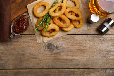 Photo of Delicious crunchy fried onion rings, sauce and beer on wooden table, flat lay. Space for text