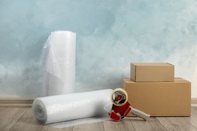 Photo of Bubble wrap rolls, tape dispenser and cardboard boxes on floor near light blue wall